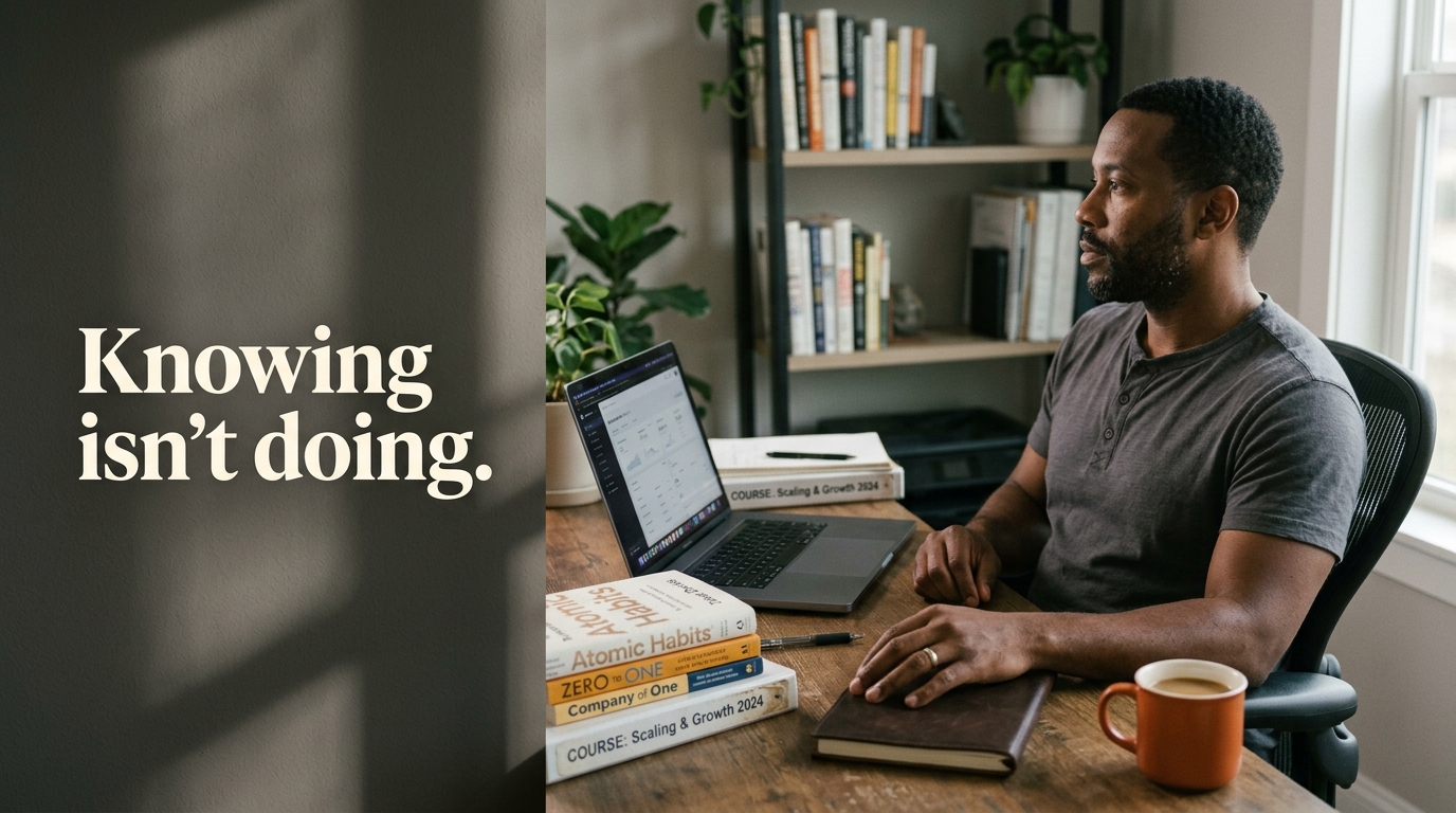 A solo business owner sits at a desk stacked with business books and course binders, looking past his open laptop, showing the knowing-doing gap between having the information and acting on it. Image text: "Knowing isn't doing."