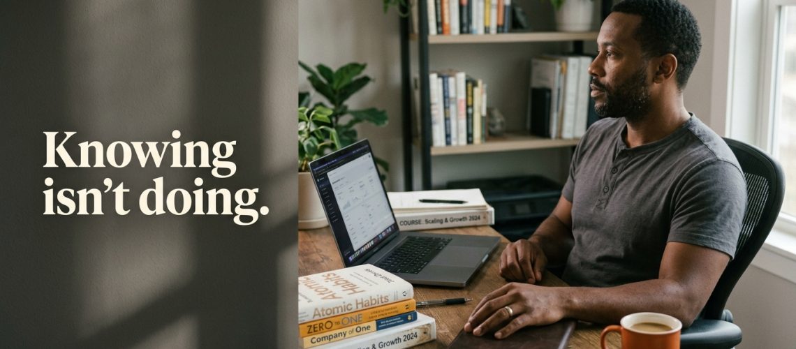 A solo business owner sits at a desk stacked with business books and course binders, looking past his open laptop, showing the knowing-doing gap between having the information and acting on it. Image text: "Knowing isn't doing."